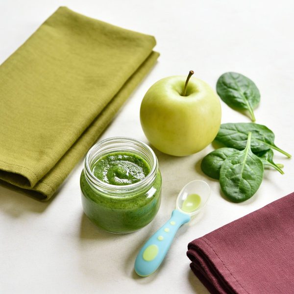 A small glass jar of smooth green spinach and apple puree with a silicone baby spoon, a green apple and fresh spinach leaves beside it.