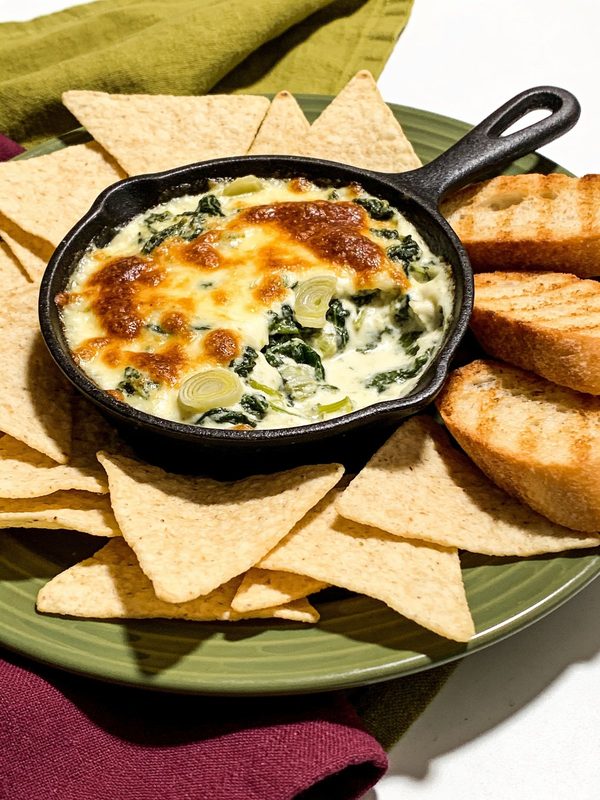 Hot spinach and artichoke dip with a browned cheese top in a small cast iron skillet, surrounded by tortilla chips and toasted baguette slices.