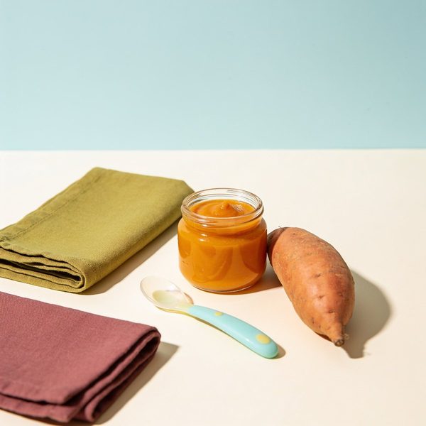 A small glass jar of smooth orange sweet potato puree with a silicone baby spoon and a whole sweet potato beside it.