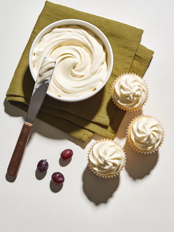 Buttercream de merengue suizo color marfil, sedoso y con remolinos suaves en un tazón, con cupcakes decorados al lado.