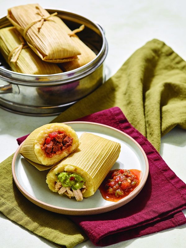 A plate with two tamales cut open, one filled with red chile pork and one with green salsa chicken, next to tomato salsa and a steamer pot of tied tamales in corn husks.