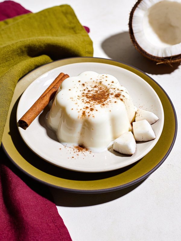 A flower-shaped coconut pudding dusted with cinnamon on a plate, with a cinnamon stick and pieces of fresh coconut.