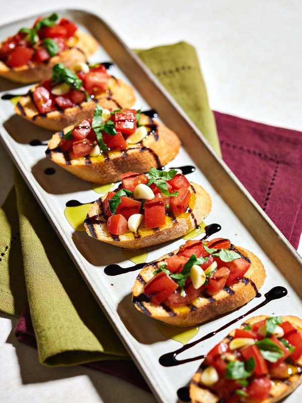 Grill-marked baguette toasts topped with diced tomatoes, basil and garlic in olive oil, drizzled with balsamic glaze on a long platter.