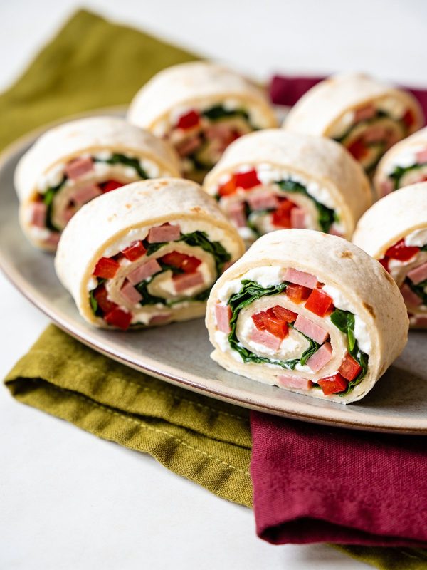 Spiral slices of flour tortilla rolled with cream cheese, ham, spinach and red bell pepper, cut side up on a platter.
