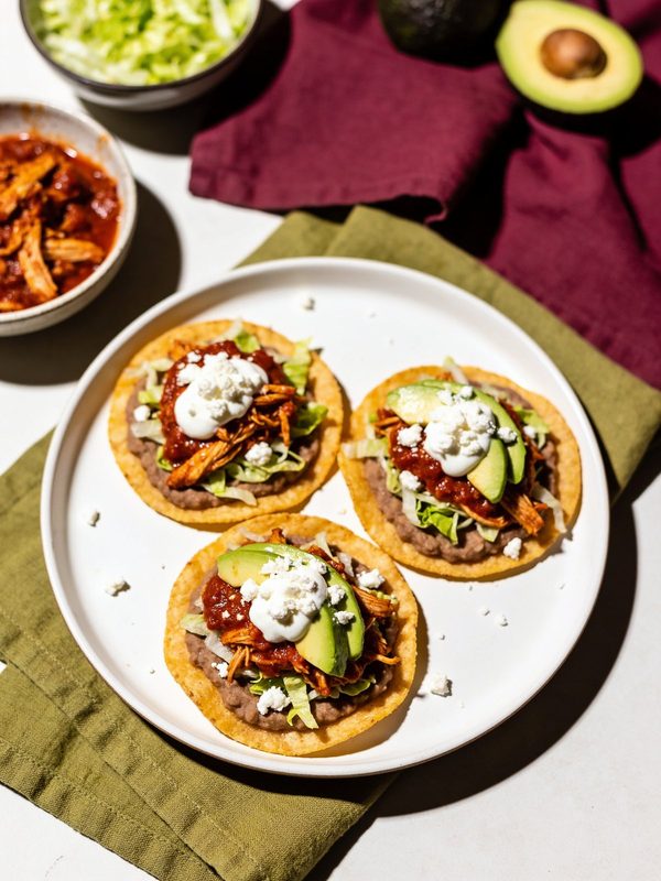 Three crisp tostadas topped with refried beans, lettuce, red chipotle chicken tinga, crema, crumbled queso fresco and avocado slices.