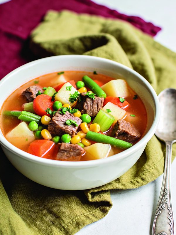 A bowl of light tomato broth with tender beef cubes, diced potatoes, carrots, green beans, corn, peas and celery, garnished with chopped parsley.
