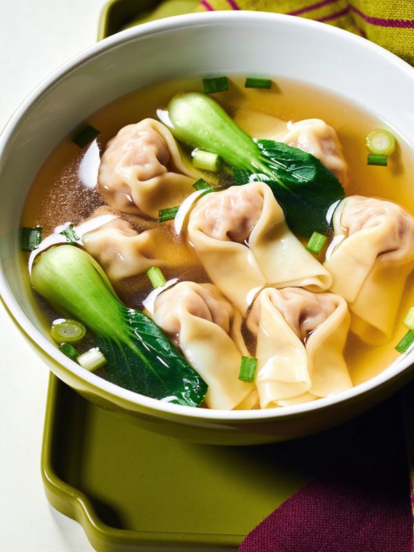 A bowl of clear light golden broth with silky pork wontons, halved baby bok choy and sliced green onions.
