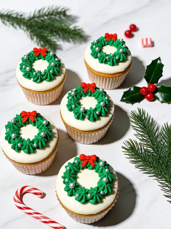 Vanilla cupcakes with smooth white frosting and piped green buttercream wreaths with silver sugar pearls and small red bows.